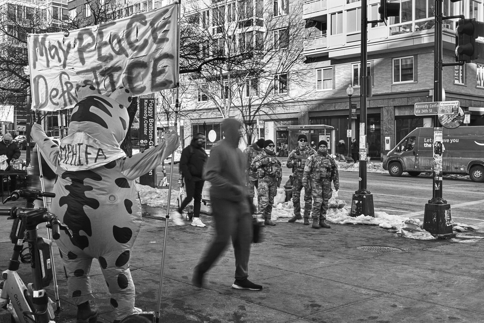 A person in a frog suit holds a sign that reads "May peace defrost ice" while National Guardsmen look on in the background