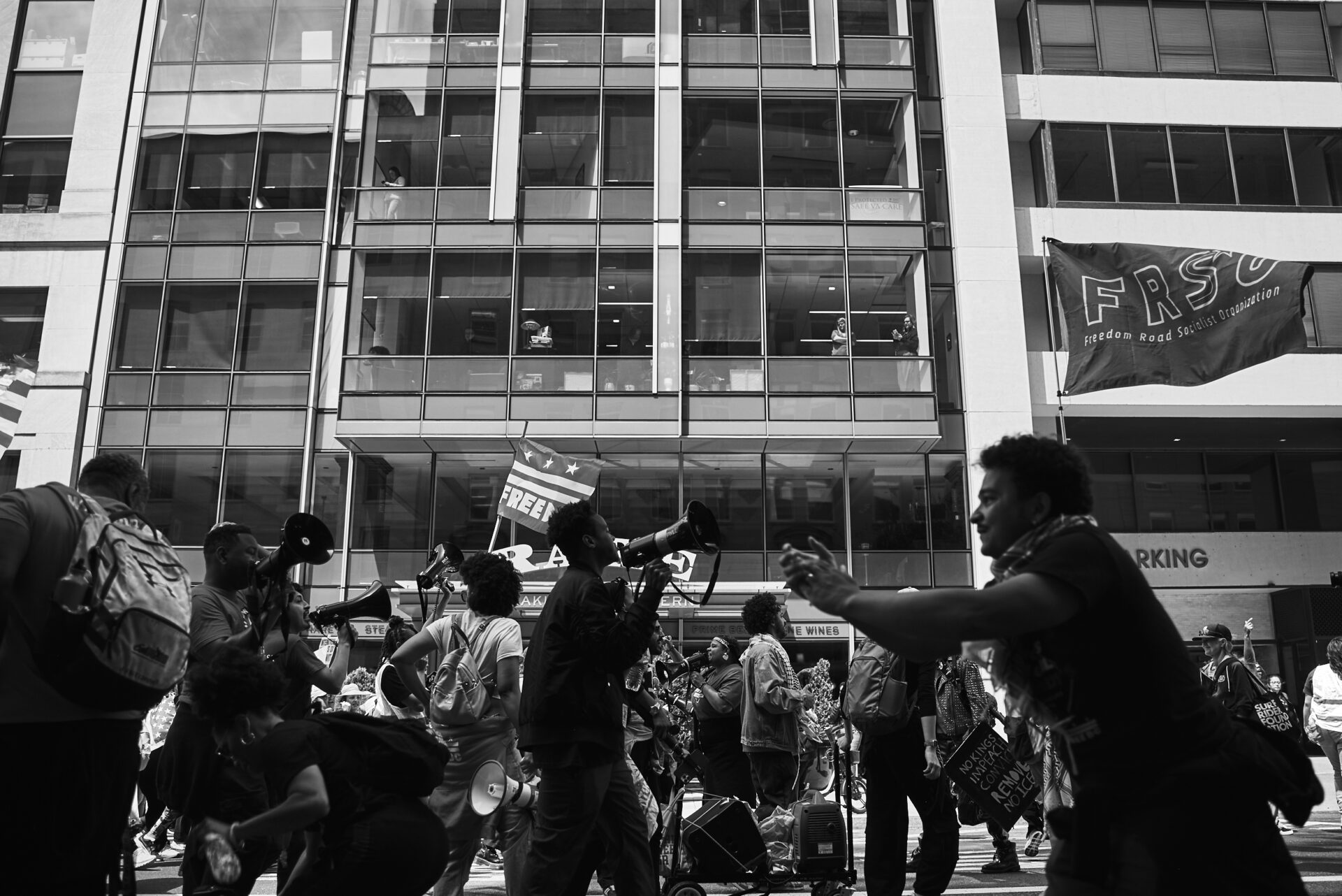 People in an office building look on and record with their phones as the protest passes through the street